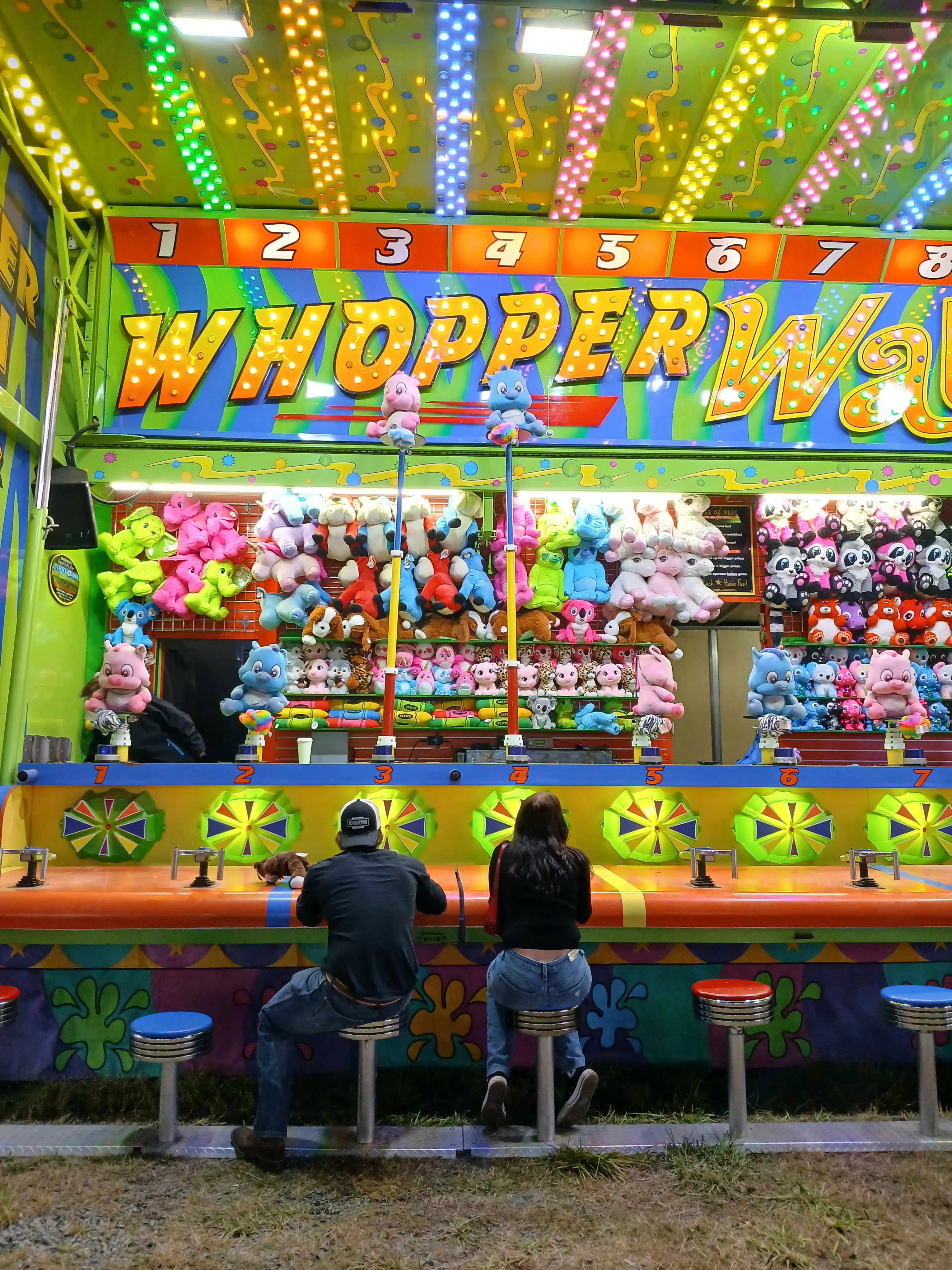 A couple sitting on stools playing a colorful carnival water game called Whopper Way at night, surrounded by vibrant lights and rows of plush toys.